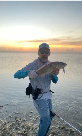 Angler holding a fish on a shallow saltwater flat at sunset, fly rod in hand